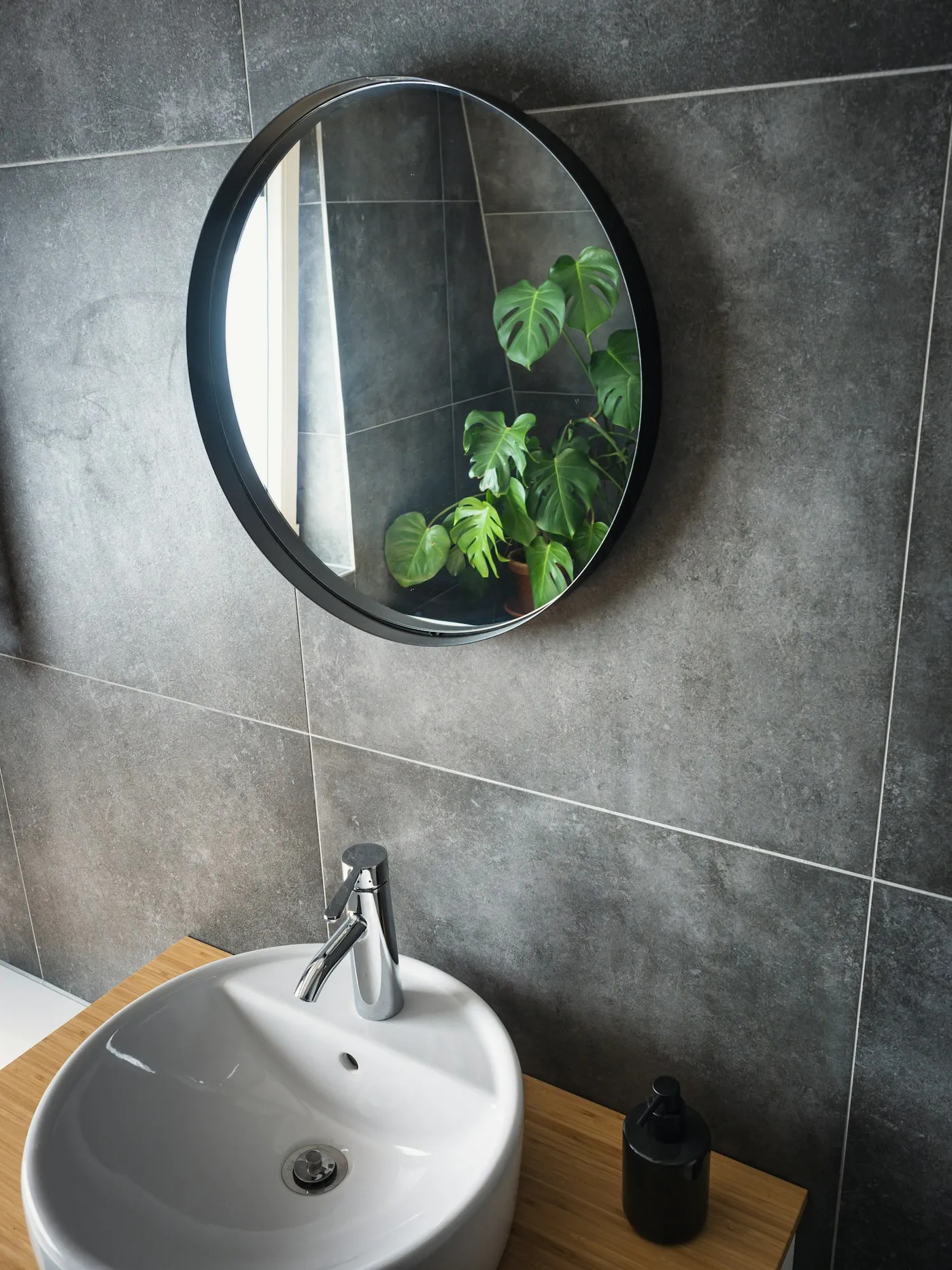 Round black-framed mirror above a sink reflecting a monstera plant.